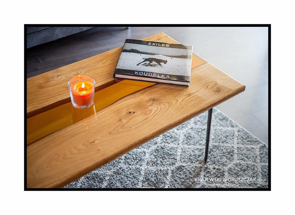 Stolik kawowy z drewna dębowego z żywicą epoksydową w kolorze bursztynowym, na metalowych nogach, dekorowany świecą i książką 'Exiles' Koudelki, na tle dywanu.