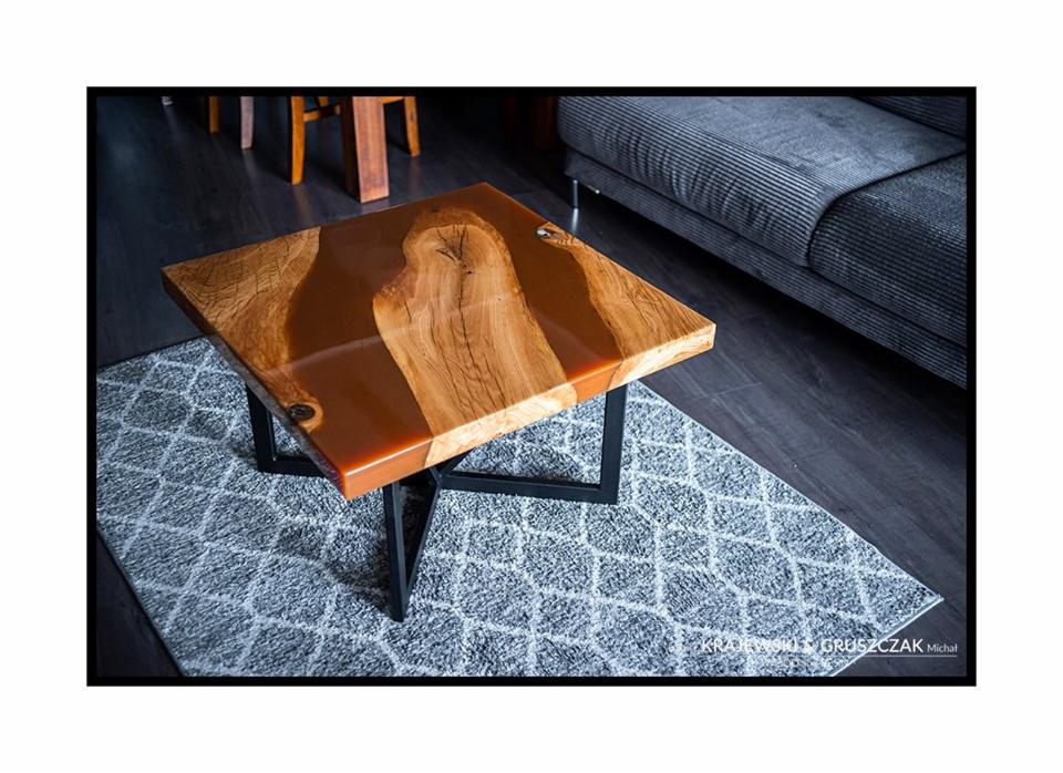 Kwadratowy stolik kawowy z blatem z drewna i żywicy epoksydowej w odcieniach brązu, na czarnych, metalowych nogach, ustawiony na szarym dywanie w geometryczny wzór. W tle widoczny fragment szarej...