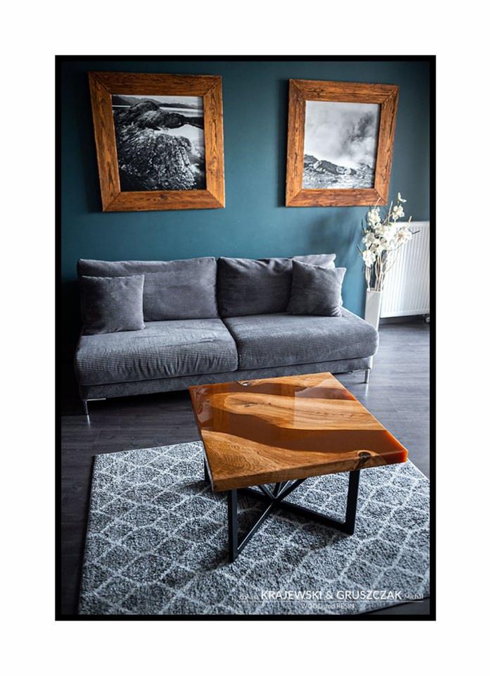 Stolik kawowy z blatem z drewna i żywicy epoksydowej na metalowej podstawie krzyżakowej, ustawiony na wzorzystym dywanie w salonie z szarą sofą i obrazami w drewnianych ramach na ścianie.