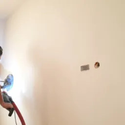 Pracownik w słuchawkach szlifuje ścianę gładziarką elektryczną z odkurzaczem, przygotowując ją do malowania; widoczne puszki instalacyjne w ścianie.