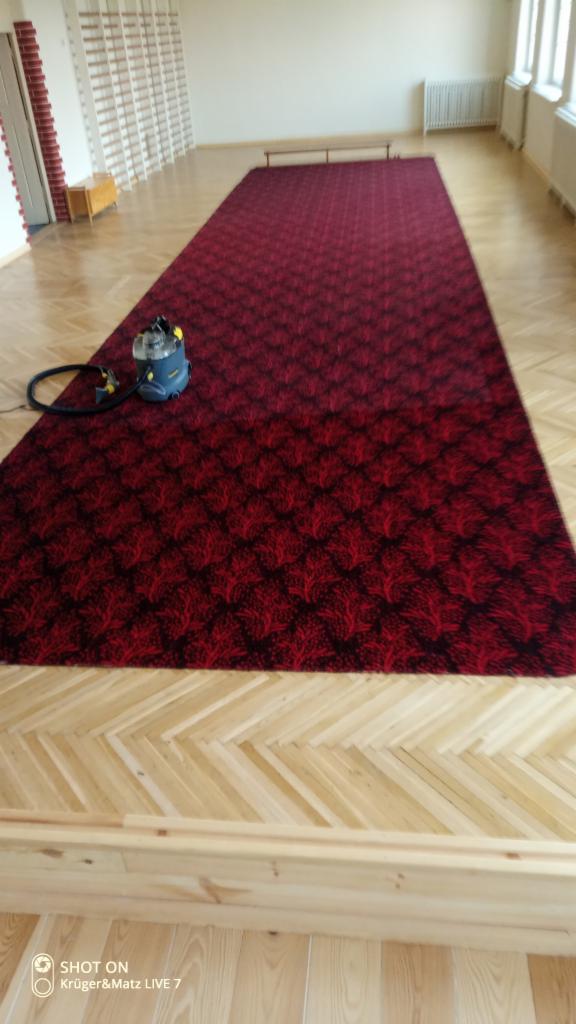 Długi, czerwony dywan z wzorem czyszczony odkurzaczem piorącym w jasnym pomieszczeniu z parkietem ułożonym w jodełkę.