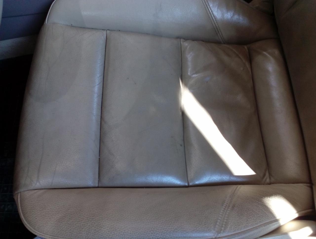 Beżowe skórzane siedzenie samochodowe z widocznymi śladami użytkowania i plamami, oświetlone ostrym promieniem słonecznym.