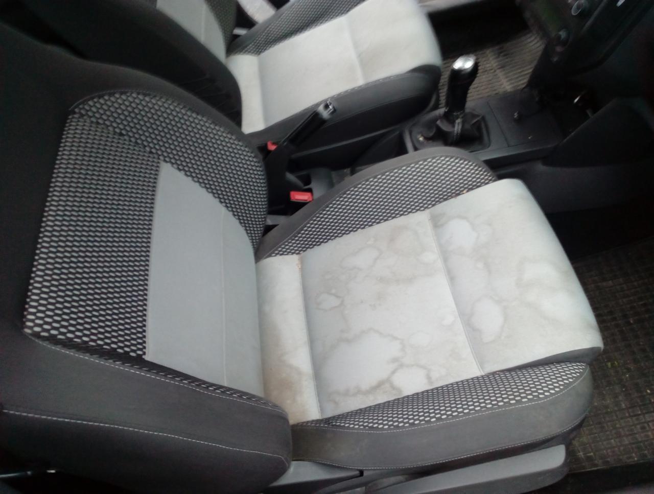 Brudne, jasne siedzenie samochodowe z plamami przed czyszczeniem, widoczna dźwignia hamulca ręcznego i lewarek zmiany biegów.