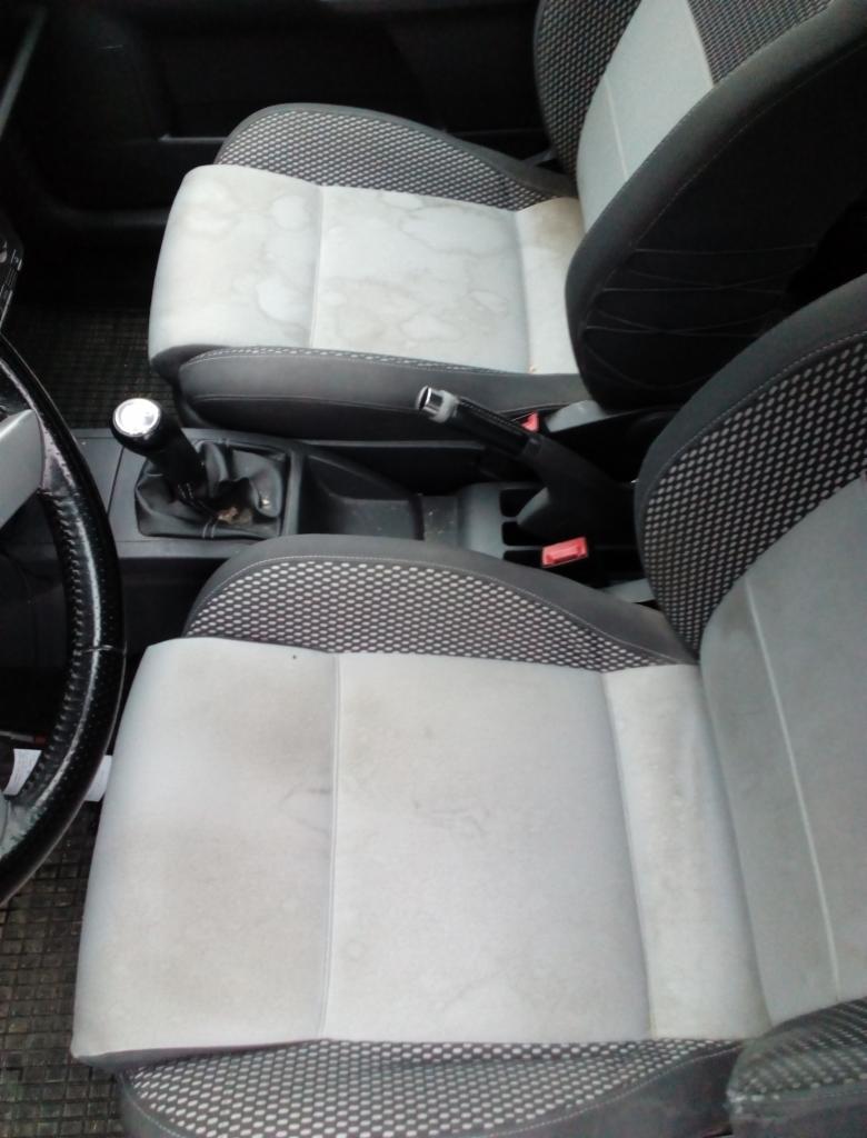 Wnętrze samochodu z widocznymi, zabrudzonymi jasnymi siedzeniami z materiałową tapicerką w szaro-czarną kratkę, obok dźwignia zmiany biegów.