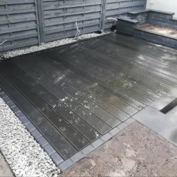 Nowoczesny taras kompozytowy w kolorze grafitowym, obrzeża z kostki, otoczony białym grysem, z szarą drewnianą ścianą w tle i schodami z szarych bloków.