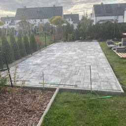 Pan Bruk Patryk Nogal - Ułożona szara kostka brukowa na posesji, widoczne ogrodzenie, trawa i domy w tle. Częściowo wykończona nawierzchnia z kostki.