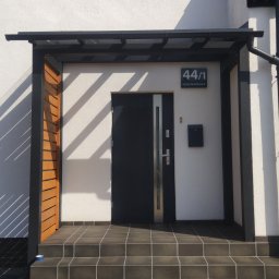 Pan Bruk Patryk Nogal - Nowoczesne wejście do domu z zadaszeniem, drewnianymi elementami i ciemną kostką na schodach. Widoczny numer domu i skrzynka pocztowa.