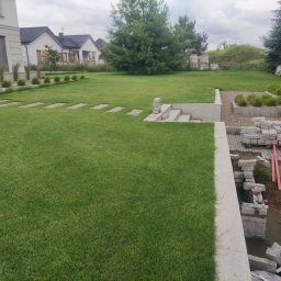 Pan Bruk Patryk Nogal - Rozległy, zielony trawnik z elementami granitowej ścieżki i schodów, w tle domy jednorodzinne i drzewa. Częściowo widoczne prace brukarskie w prawym dolnym rogu.