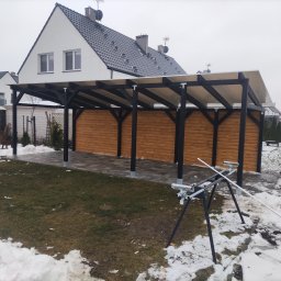 Pan Bruk Patryk Nogal - Zadaszenie tarasu z drewnianą zabudową, czarne słupy, jasna plandeka, kostka brukowa, dom w tle, resztki śniegu. Widoczne narzędzia montażowe.