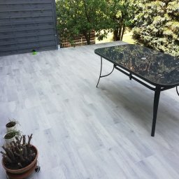 Pan Bruk Patryk Nogal - Jasnoszary taras z płyt imitujących drewno, czarny stół z szklanym blatem, doniczki z roślinami, szara drewniana ściana i zieleń drzew w tle.