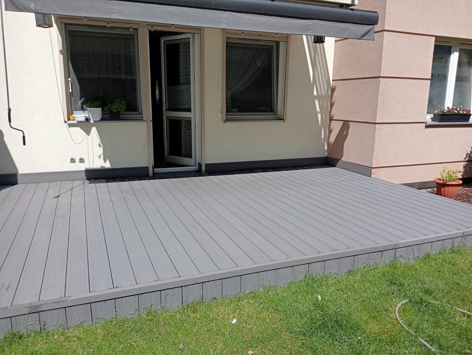 Szary taras kompozytowy przylegający do domu z otwartymi drzwiami balkonowymi, widok z poziomu trawnika. Widoczne detale wykończenia i rośliny w donicach.
