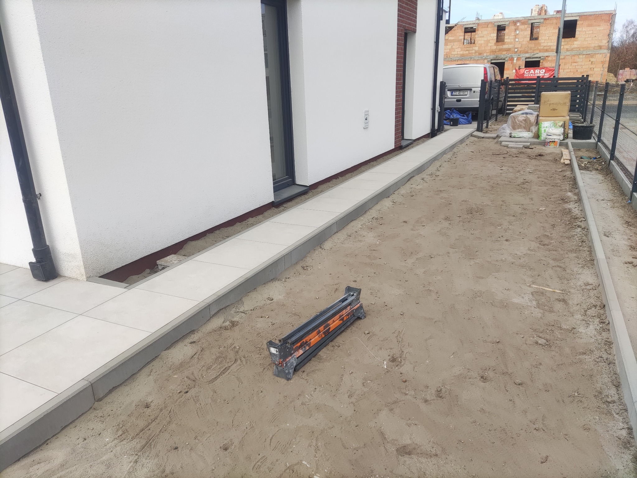 Ścieżka z jasnych płyt ułożona wzdłuż białej ściany budynku, z widocznym narzędziem do cięcia płytek na piaszczystym podłożu. W tle budowa i samochód dostawczy.