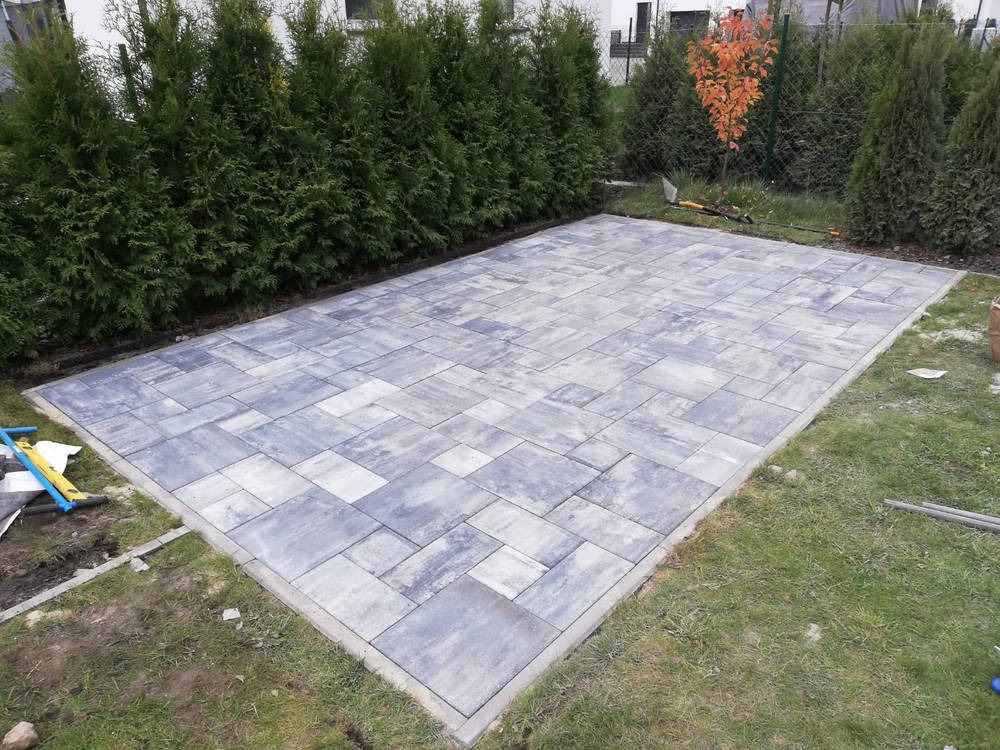 Ułożona, prostokątna nawierzchnia z szarej kostki brukowej na trawniku, obrzeża betonowe, w tle rząd tuj i drzewko z pomarańczowymi liśćmi.