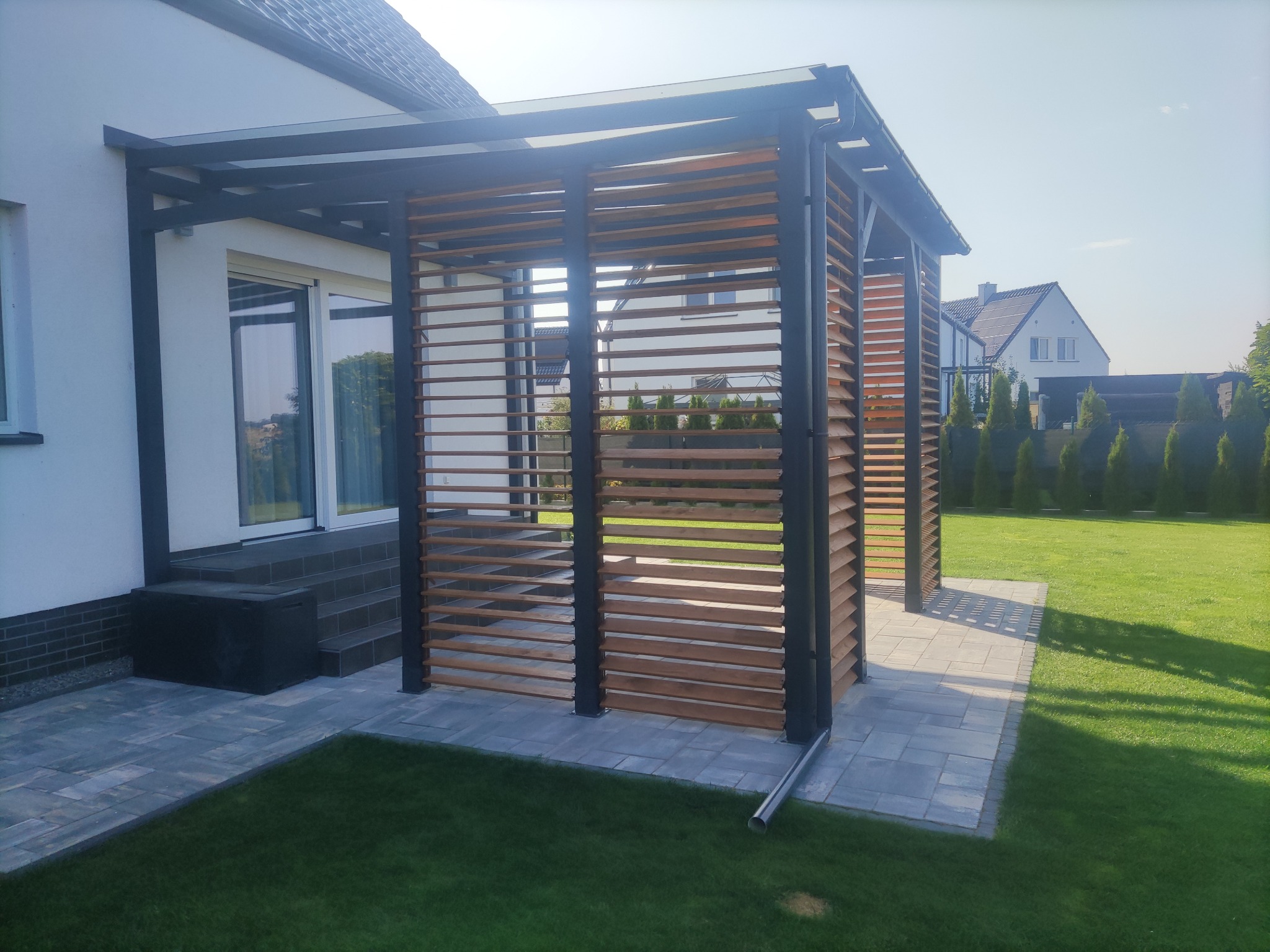 Nowoczesna pergola z drewnianymi lamelami i metalową konstrukcją, przylegająca do domu, z kostką brukową i trawnikiem. Widok na sąsiedni dom i tuje.
