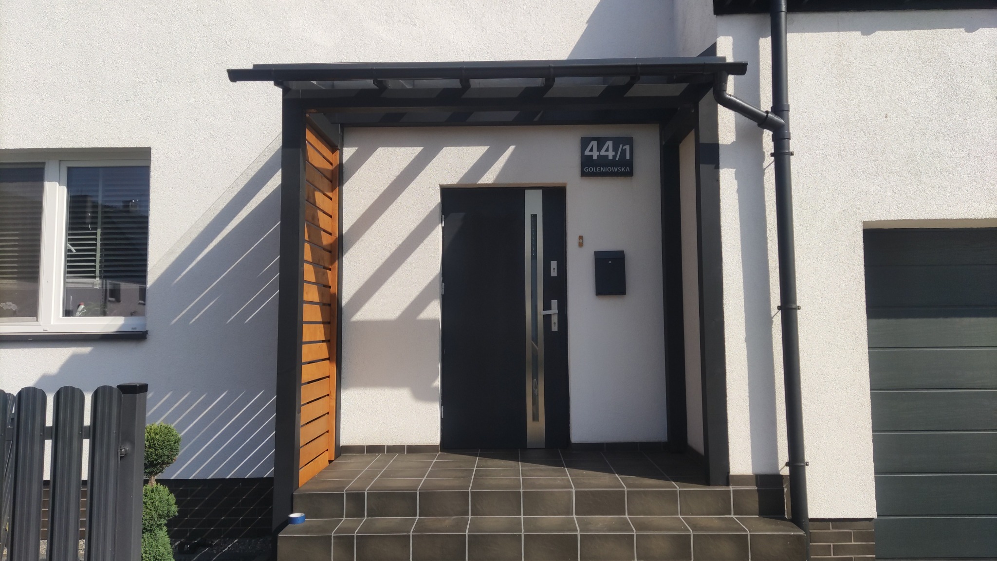 Nowoczesne wejście do domu z zadaszeniem, drewnianymi elementami i ciemną kostką na schodach. Widoczny numer domu i skrzynka pocztowa.