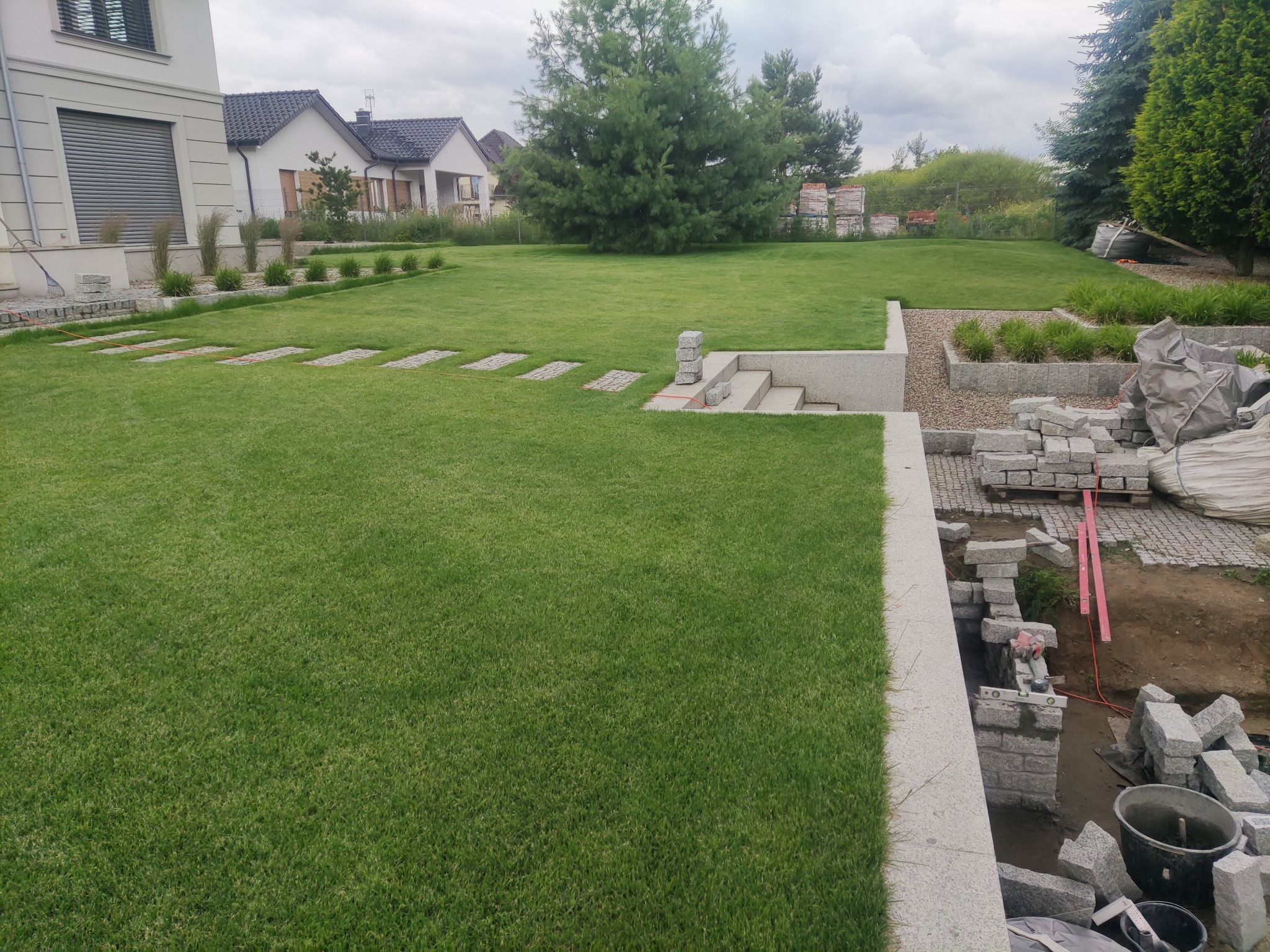 Rozległy, zielony trawnik z elementami granitowej ścieżki i schodów, w tle domy jednorodzinne i drzewa. Częściowo widoczne prace brukarskie w prawym dolnym rogu.