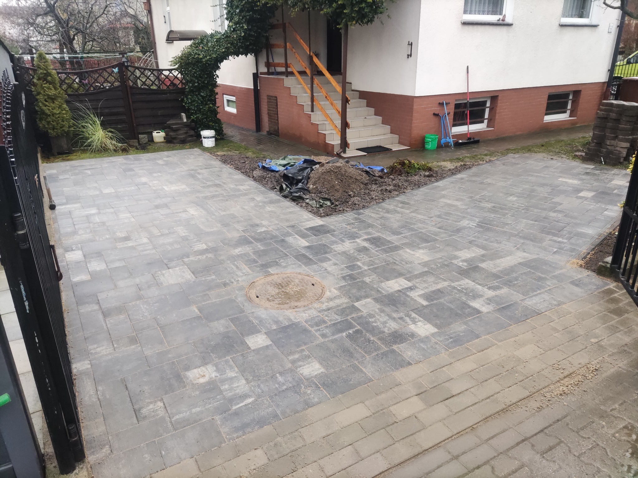 Ułożona szara kostka brukowa na podjeździe przed domem z zielonymi akcentami roślinności i elementami wyposażenia. Widoczny właz kanalizacyjny.