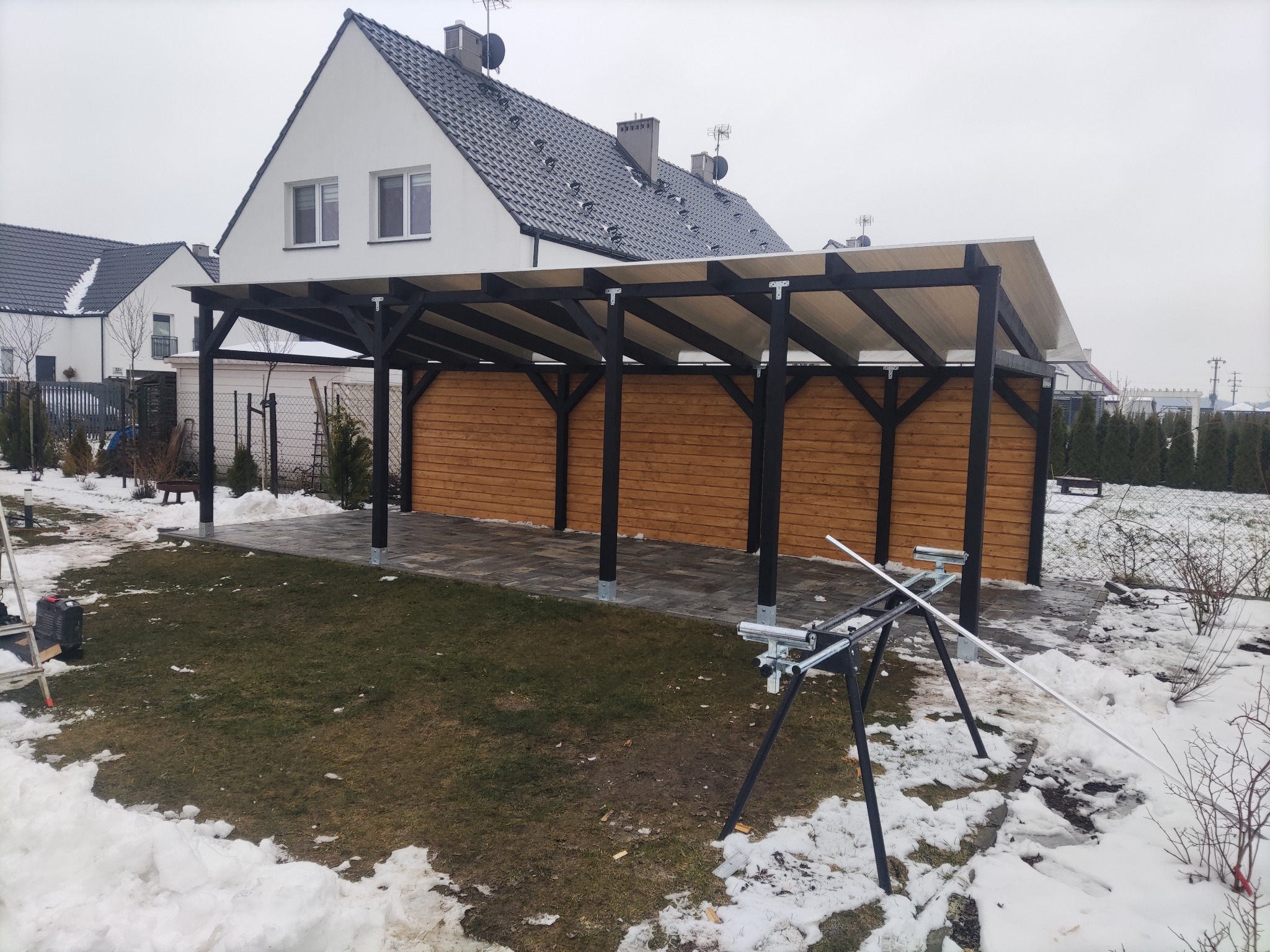 Zadaszenie tarasu z drewnianą zabudową, czarne słupy, jasna plandeka, kostka brukowa, dom w tle, resztki śniegu. Widoczne narzędzia montażowe.