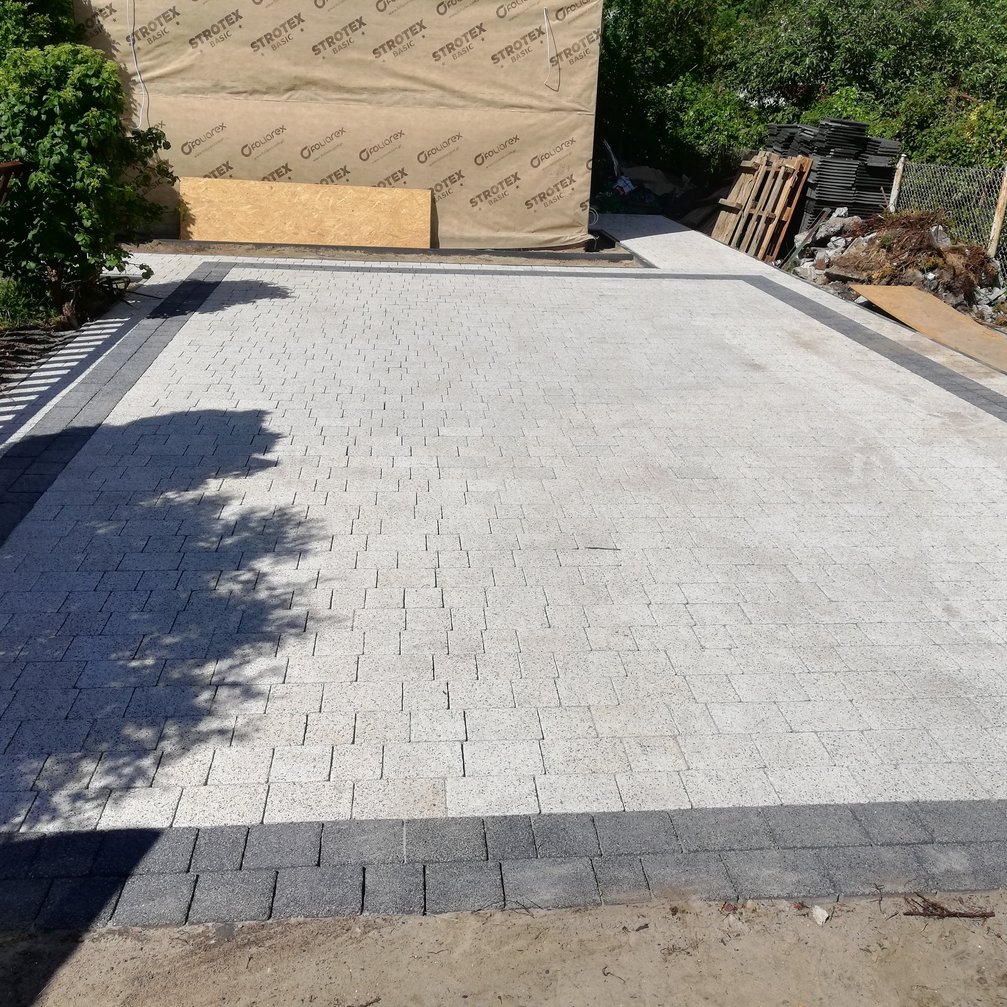 Jasnoszara kostka brukowa ułożona na dużej powierzchni, obramowana ciemniejszą kostką. W tle widoczne materiały budowlane i zieleń.