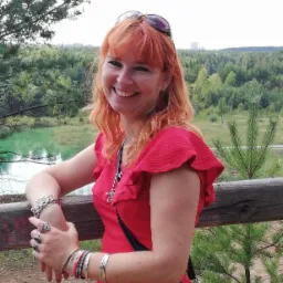 Uśmiechnięta kobieta z rudymi włosami w czerwonej sukience opiera się o drewniane ogrodzenie z widokiem na jezioro i las w tle.