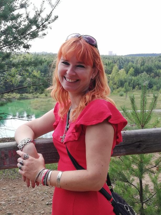 Uśmiechnięta kobieta z rudymi włosami w czerwonej sukience opiera się o drewniane ogrodzenie z widokiem na jezioro i las w tle.