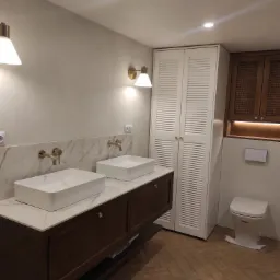 Nowoczesna łazienka z dwoma białymi umywalkami nablatowymi, złotą armaturą, marmurowym blatem na szafce z ciemnego drewna, białą szafą z lamelami i drewnianą szafką wiszącą z plecionką.