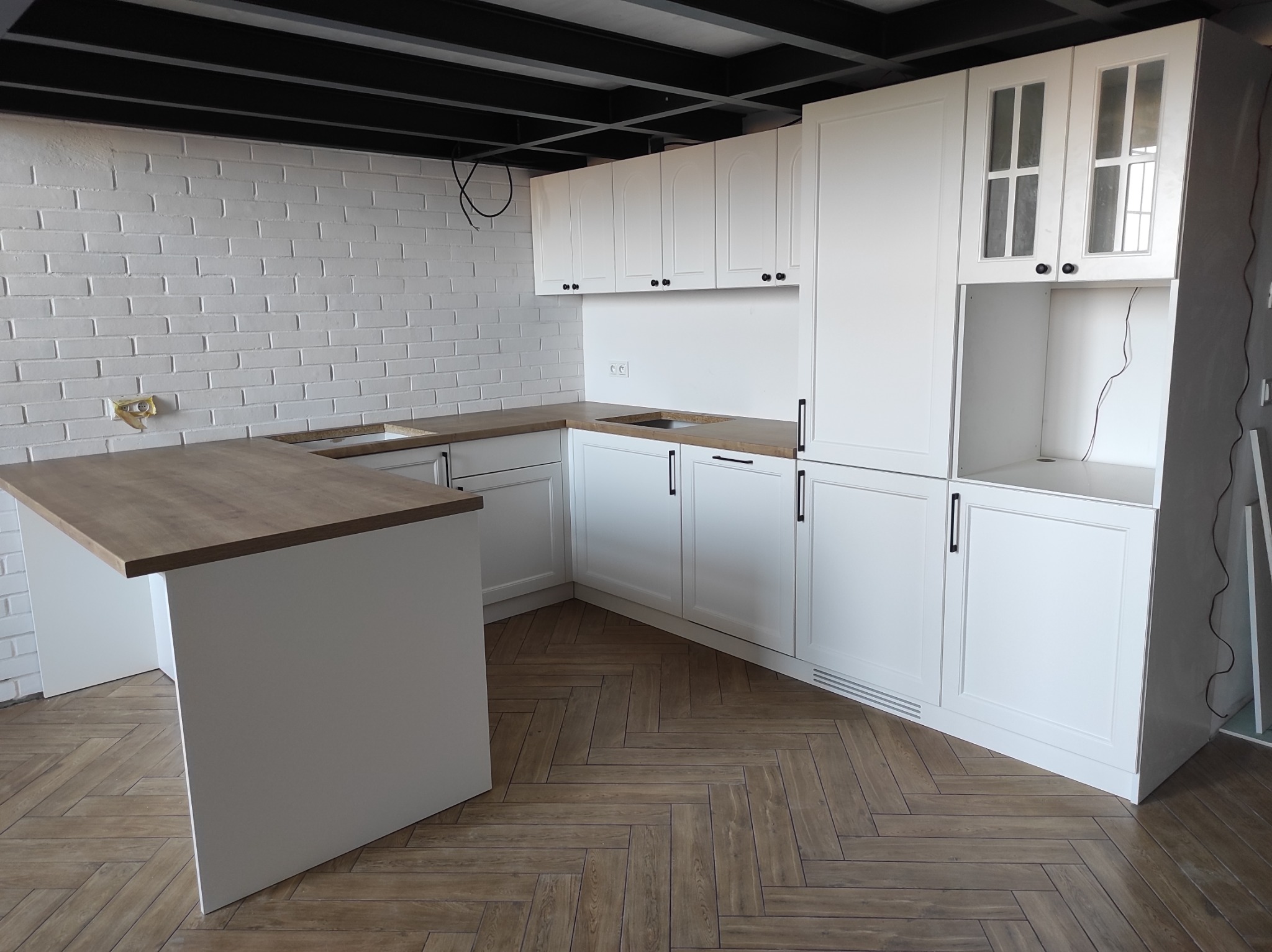 Biała kuchnia z drewnianym blatem i wyspą, widok na ceglaną ścianę i parkiet w jodełkę.