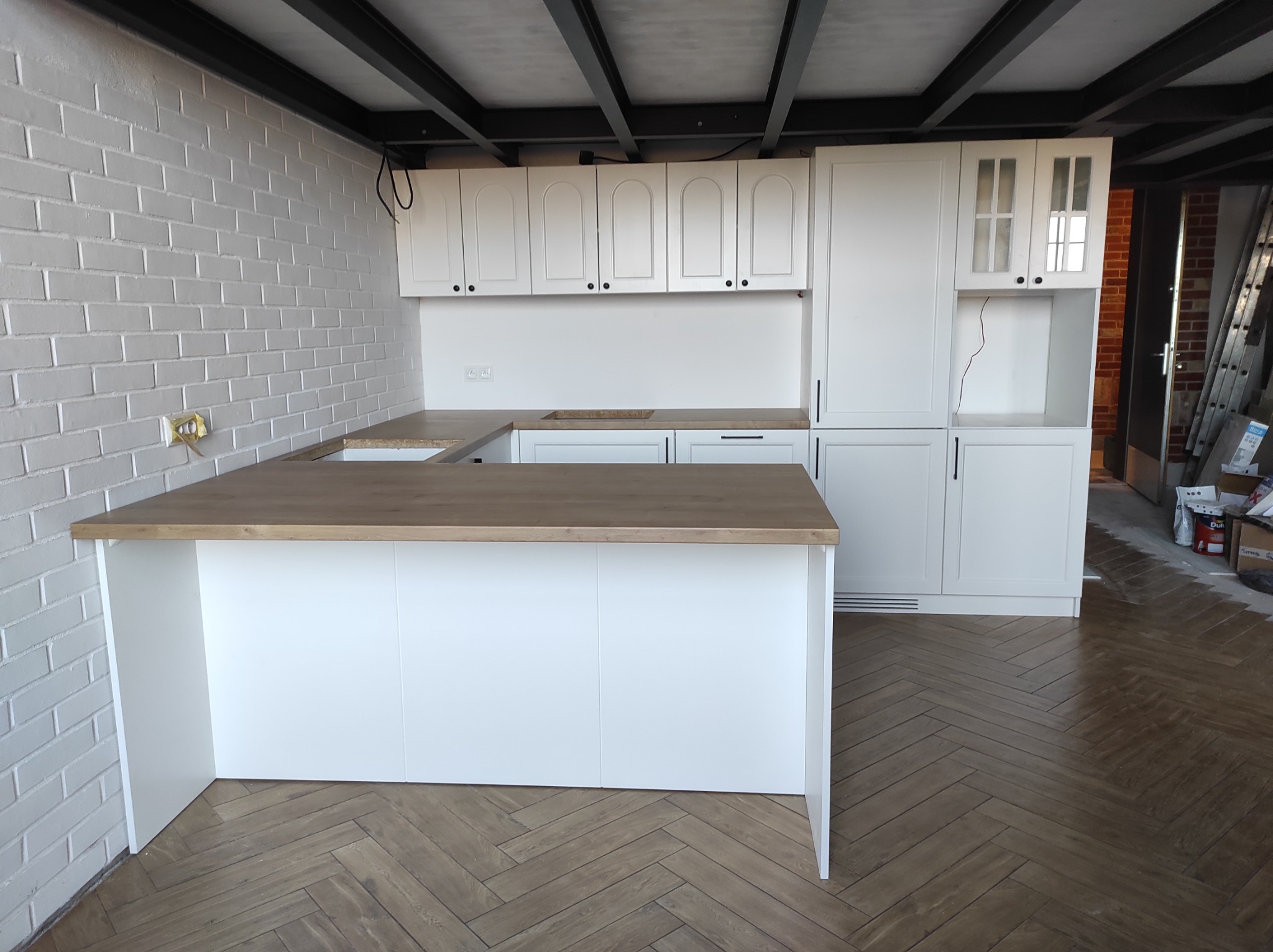 Nowoczesna biała kuchnia z drewnianym blatem i ceglaną ścianą, widoczne elementy wykończeniowe i podłoga ułożona w jodełkę.