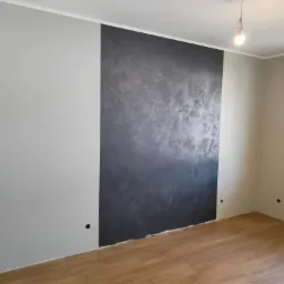 Pokój po remoncie: ściany pomalowane na szaro z dekoracyjnym panelem w odcieniach szarości, drewniana podłoga, widoczne gniazdka elektryczne bez osprzętu.