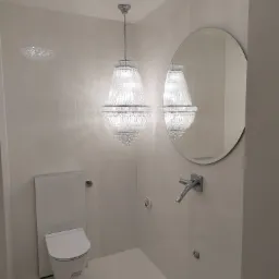 Nowoczesna łazienka z wiszącą toaletą Geberit, okrągłym lustrem i kryształowym żyrandolem, ściany wykończone panelami w odcieniach bieli.