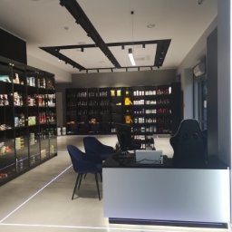 Refresh - Nowoczesne wnętrze biura z czarnymi regałami wypełnionymi produktami, minimalistycznym oświetleniem i recepcją z niebieskimi krzesłami.