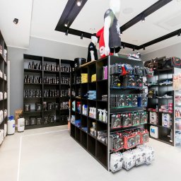 Refresh - Wnętrze sklepu z akcesoriami do auto detailingu, z czarnymi regałami wypełnionymi butelkami, opakowaniami i narzędziami, manekiny w koszulkach i czapkach na górze regałów, jasna podłoga...