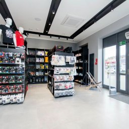 Refresh - Wnętrze sklepu z artykułami do detailingu samochodowego, regały z produktami, manekiny w koszulkach, jasna podłoga i szare ściany, widoczne drzwi wejściowe.