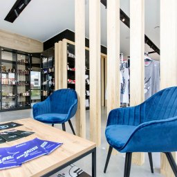 Refresh - Jasne wnętrze biura z niebieskimi fotelami welurowymi i drewnianym stołem, za którym znajdują się przeszklone regały z produktami i drewniane panele działowe.