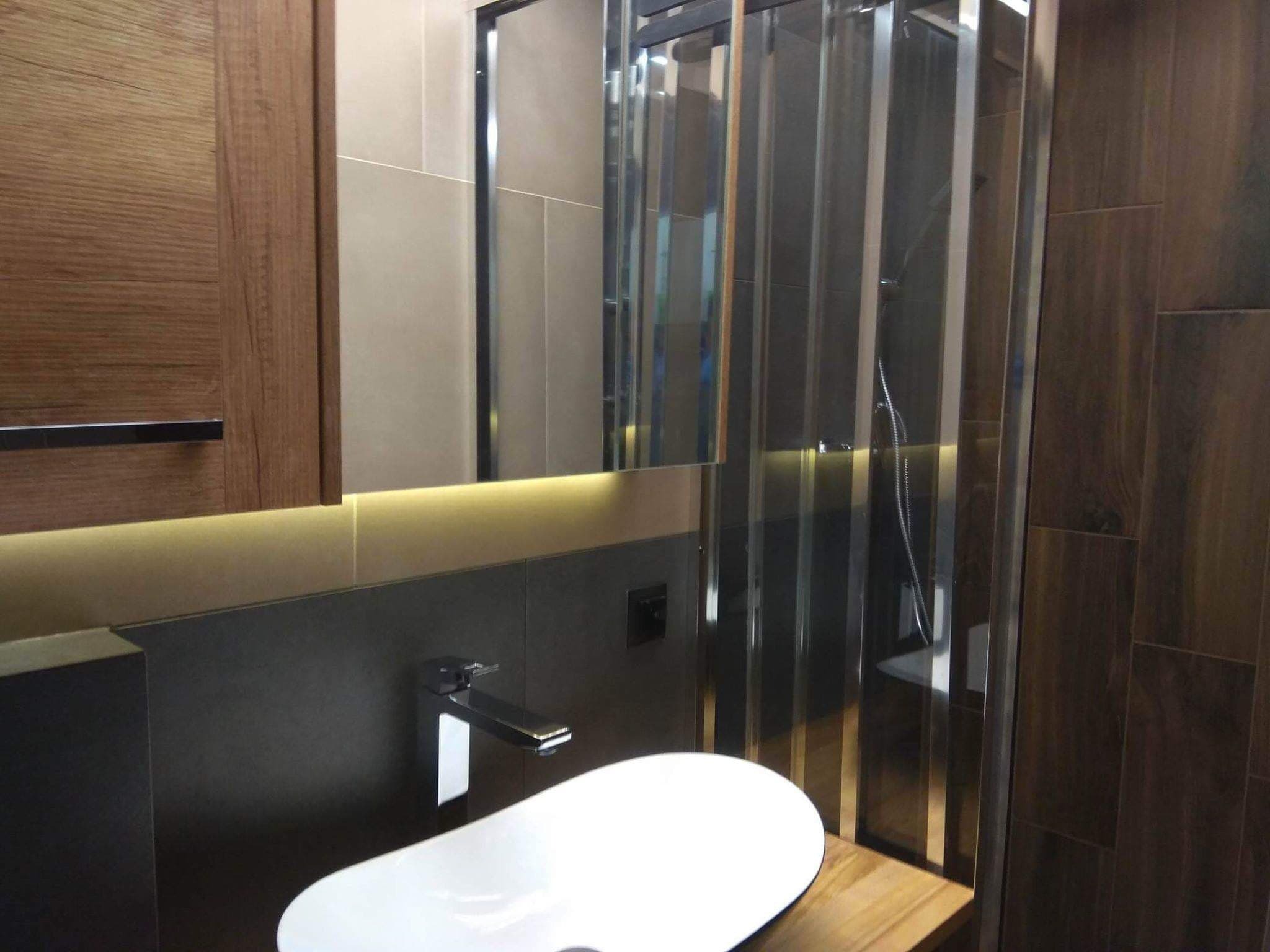 Nowoczesna łazienka z umywalką nablatową, baterią w kolorze czarnym i kabiną prysznicową ze szklanymi drzwiami, oświetleniem LED pod szafką i drewnianymi elementami wykończenia.