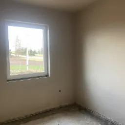 Pokój po tynkowaniu maszynowym, ściany gładkie, surowe, w rogu widoczne rury grzewcze przy podłodze, okno z widokiem na zewnątrz.