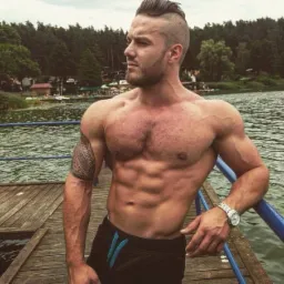 Mężczyzna o muskularnej sylwetce z tatuażem na ramieniu, oparty o barierkę pomostu nad jeziorem, prezentujący formę fizyczną.