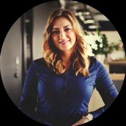 Uśmiechnięta kobieta z brązowymi włosami w niebieskiej koszuli, pozująca w biurze z roślinami w tle.