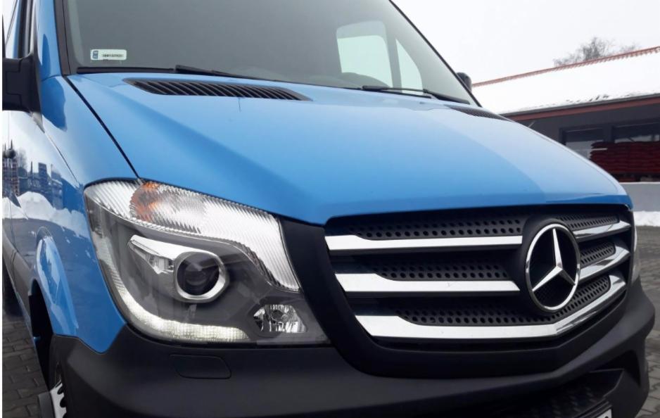 Niebieski samochód dostawczy Mercedes-Benz Sprinter z widocznym przodem, reflektorem i grillem z logo, zaparkowany na zewnątrz w zimowy dzień.