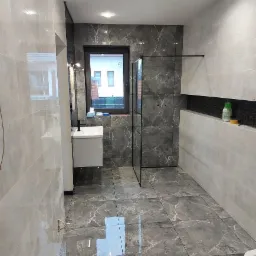 Wykończona łazienka z szarymi płytkami imitującymi marmur na ścianach i podłodze, kabina prysznicowa typu walk-in z czarnymi profilami, biała szafka z umywalką i czarną baterią, okno z widokiem...