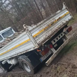 Biały, lekko zabłocony samochód ciężarowy z żółtymi pasami, wypełniony pociętymi gałęziami, stoi na błotnistej drodze w lesie.