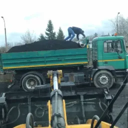 Operator równa powierzchnię czarnego materiału w skrzyni zielonej ciężarówki, widok z kabiny żółtej ładowarki teleskopowej, pochmurne niebo w tle.