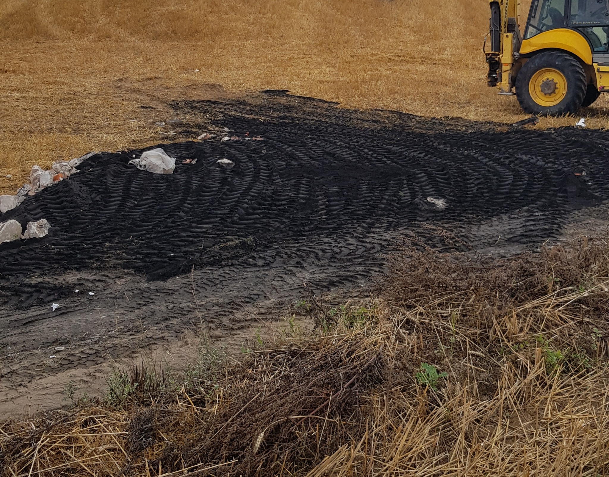 Żółty traktor częściowo widoczny na skraju pola w Kartuzach, obok świeżo wyrównanego, czarnego terenu z widocznymi śladami opon i gruzem.