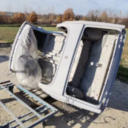 Podwozie samochodu po zabezpieczeniu antykorozyjnym i wstępnym malowaniu, umieszczone na metalowym stelażu na zewnątrz, z częściowo zakrytą folią ochronną i widocznym tłem polnym z drzewami.