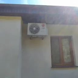 Biała jednostka zewnętrzna klimatyzacji LG Dual Inverter zamontowana na jasnej ścianie budynku pomiędzy dwoma oknami z brązowymi ramami, w tle widoczne drzewa w słoneczny dzień.