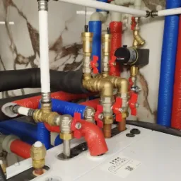 Instalacja hydrauliczna z rurami w kolorach czerwonym i niebieskim oraz mosiężnymi zaworami z czerwonymi pokrętłami, widoczny fragment kotła grzewczego z naklejką informacyjną.