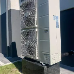 Zewnętrzna jednostka pompy ciepła Buderus z dwoma wentylatorami, umieszczona na ciemnym, granitowym cokole przed budynkiem z szarą elewacją.