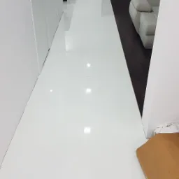 Biała, błyszcząca podłoga z płytek w nowoczesnym wnętrzu z widocznym fragmentem beżowego dywanu i szarą sofą w tle.