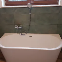 Nowoczesna, wolnostojąca wanna Roca z chromowaną baterią i słuchawką prysznicową na tle oliwkowo-zielonych płytek w łazience z oknem.