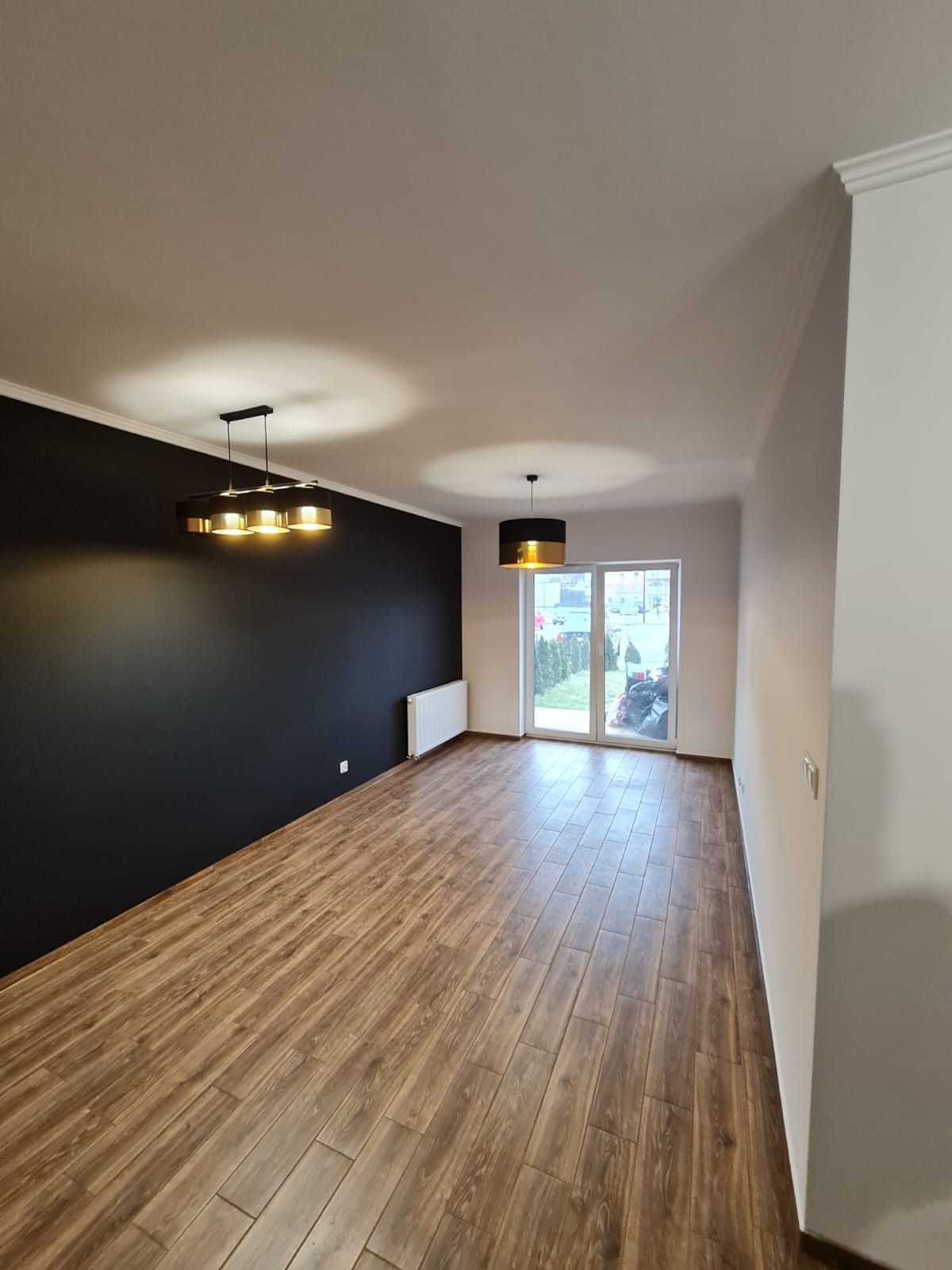 Wykończone wnętrze w Łodzi z drewnopodobną podłogą, pomalowaną na czarno ścianą z grzejnikiem i nowoczesnym oświetleniem sufitowym.